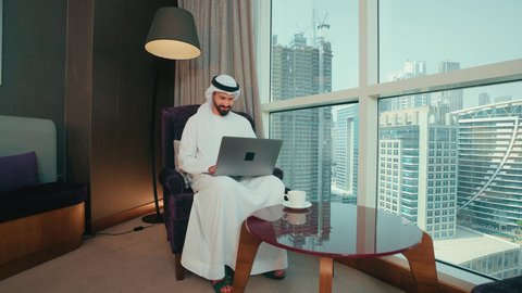 العمل عن بعد، مهنة ووظيفة مكتبية، استخدام التقنية الحديثه المتطورة في الحياة العملية، ايماءات الاندماج والتركيز، رجل عربي خليجي اماراتي يرتدي الكندورة و الغتره يجلس على الكرسي ويعمل على الحاسب المحمول في صالة المنزل، النظر الى شي ما