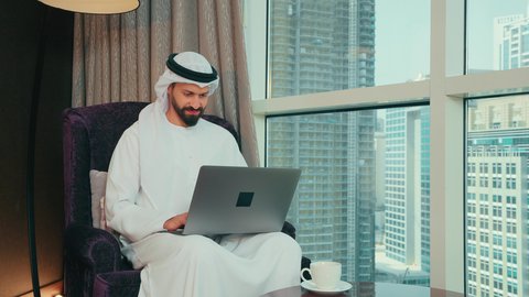 ايماءات الاندماج والتركيز، العمل عن بعد، مهنة ووظيفة مكتبية، استخدام التقنية الحديثه المتطورة في الحياة العملية، رجل عربي خليجي اماراتي يرتدي الكندورة و الغتره يجلس على الكرسي ويعمل على الحاسب المحمول في صالة المنزل، النظر الى شي ما