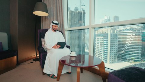 استخدام تطبيقات وبرامج الأجهزة اللوحية، النظر الى شيء ما، انجاز مهام العمل عبر الانترنت، استعمال التكنولوجيا والتقنية الحديثة، المحادثة مع الأهل والأصدقاء، رجل عربي خليجي اماراتي يرتدي الكندورة و الغتره يجلس على كرسي بجانب النافذة يحمل بيده جهاز التابلت