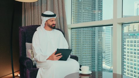 Looking at something, completing work tasks online, using modern technology and techniques, chatting with family and friends, an Arab Gulf Emirati man wearing a kandura and ghutrah sitting on a chair by the window holding a tablet in his hand.