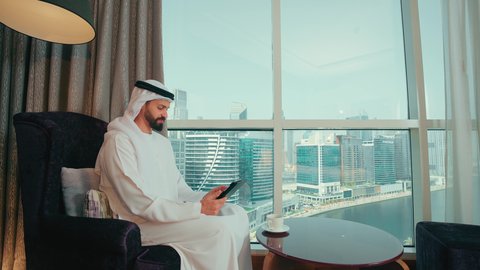 Browsing social media sites on a tablet, using modern technology and techniques, chatting with family and friends, a side shot of an Arab Gulf Emirati man wearing a kandura and ghutrah sitting on a chair by the window holding a tablet in his hand, looking at something.
