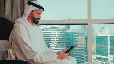 استعمال التكنولوجيا والتقنية الحديثة, تصفح مواقع التواصل الاجتماعي عبر الجهاز اللوحي، المحادثة مع الأهل والأصدقاء،تصوير من الجانب لرجل عربي خليجي اماراتي يرتدي الكندورة و الغتره يجلس على كرسي بجانب النافذة يحمل بيده جهاز التابلت، النظر الى شيء ما