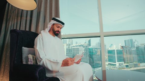 تحفيظ وتفسير كتاب الله، العبادة والتقرب إلى الله ، مفهوم الثقافة والتعلم الذاتي، ايماءات التركيز والاستمتاع, تصوير من الجانب لرجل عربي خليجي اماراتي يرتدي الكندوره والغترة  يجلس على كرسي بجانب النافذة يقوم بقراة كتاب تفسير القران