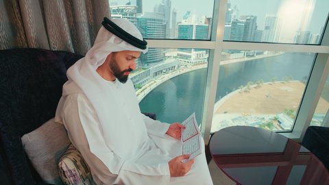 العبادة والتقرب إلى الله ، مفهوم الثقافة والتعلم الذاتي، ايماءات التركيز والاستمتاع, تحفيظ وتفسير كتاب الله, تصوير من الجانب لرجل عربي خليجي اماراتي يرتدي الكندوره والغترة  يجلس على كرسي بجانب النافذة يقوم بقراة كتاب تفسير القران