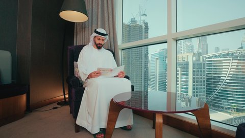 مفهوم الثقافة والتعلم الذاتي، ايماءات التركيز والاستمتاع, تحفيظ وتفسير كتاب الله, رجل عربي خليجي اماراتي يرتدي الكندوره والغترة  يجلس على كرسي بجانب النافذة يقوم بقراة كتاب تفسير القران ، مفهوم العبادة والتقرب إلى الله