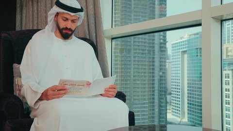 Gestures of focus and enjoyment, the concept of culture and self-learning, memorization and interpretation of the Book of God, an Arab Gulf Emirati man wearing a kandura and ghutrah sitting on a chair by the window reading a book on the interpretation of the Quran, the concept of worship and drawing closer to God.