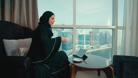 الاستمتاع بتناول كوب من القهوه، تصفح مواقع ووسائل التواصل الاجتماعي، مفهوم انجار المشاريع عن بعد، دمج التقنية في الحياة اليومية والعملية، تصوير من الجانب لامرأة عربية خليجية اماراتية ترتدي العباءة والحجاب تجلس على كرسي  تقوم بانجاز مهام العمل عبر جهاز التابلت، سيده سعودية تقوم باستخدام الاجهزة التقنية الحديثة