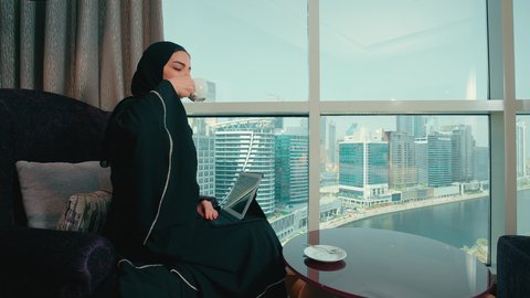 تصفح مواقع ووسائل التواصل الاجتماعي، مفهوم انجار المشاريع عن بعد، دمج التقنية في الحياة اليومية والعملية، تصوير من الجانب لامرأة عربية خليجية اماراتية ترتدي العباءة والحجاب تجلس على كرسي  تقوم بانجاز مهام العمل عبر جهاز التابلت، سيده سعودية تقوم باستخدام الاجهزة التقنية الحديثة، الاستمتاع بتناول كوب من القهوه