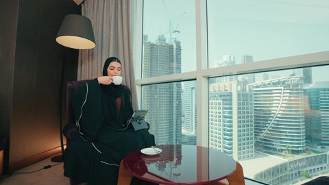 The concept of remote project completion, integrating technology into daily and professional life, an Arab Gulf Emirati woman wearing an abaya and hijab sitting on a chair completing work tasks via a tablet, a Saudi woman using modern technological devices, browsing websites and social media, enjoying a cup of coffee.