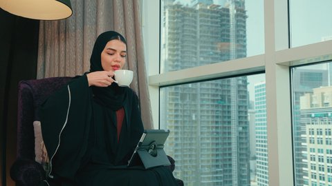 دمج التقنية في الحياة اليومية والعملية، امرأة عربية خليجية اماراتية ترتدي العباءة والحجاب تجلس على كرسي  تقوم بانجاز مهام العمل عبر جهاز التابلت،سيده سعودية تقوم باستخدام الاجهزة التقنية الحديثة ، تصفح مواقع ووسائل التواصل الاجتماعي، مفهوم انجار المشاريع عن بعد، الاستمتاع بتناول كوب من القهوه