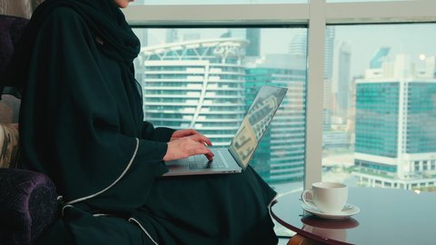 دمج التكنولوجيا بالعمل، النقر على لوحه المفتايح، ادارة الأعمال عن بعد، استخدام الأجهزة الحديثة والمتطورة، تصوير من الجانب لامراة عربية خليجية اماراتية ترتدي العباءة والحجاب تجلس على كرسي مريح تقوم باستخدام الحاسوب المحمول، سيده سعودية تقوم بإنجاز المهام المطلوبة عبر اللابتوب، مهن ووظائف نسائية