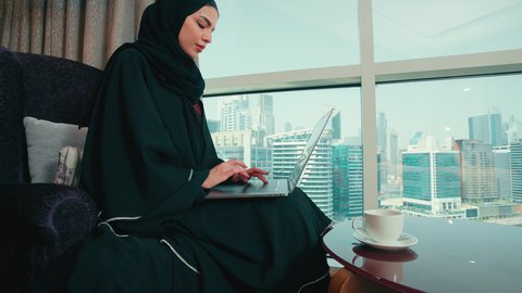 النقر على لوحه المفتايح، ادارة الأعمال عن بعد، استخدام الأجهزة الحديثة والمتطورة، تصوير من الجانب لامراة عربية خليجية اماراتية ترتدي العباءة والحجاب تجلس على كرسي مريح تقوم باستخدام الحاسوب المحمول، دمج التكنولوجيا بالعمل، سيده سعودية تقوم بإنجاز المهام المطلوبة عبر اللابتوب، مهن ووظائف نسائية