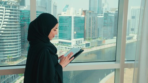 تصفح مواقع التواصل الاجتماعي، إنجاز مهام العمل عن بعد, التطبيقات التقنية للأجهزة اللوحية, النظر الى شيء ما، استخدام الأجهزة والتقنيات الحديثة والمتطورة, تصوير من الجانب لامراة عربية خليجية اماراتية ترتدي الحجاب و العباءة تقف بجانب النافذة تمسك بيدها جهاز التابليت, التسوق الالكتروني عبر الأونلاين