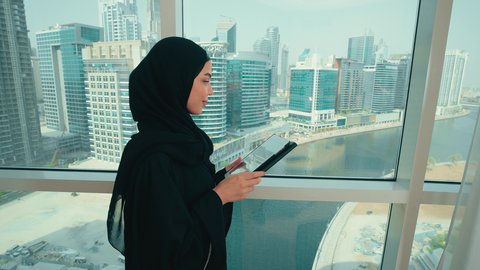 إنجاز مهام العمل عن بعد, التطبيقات التقنية للأجهزة اللوحية, النظر الى شيء ما، استخدام الأجهزة والتقنيات الحديثة والمتطورة, تصوير من الجانب لامراة عربية خليجية اماراتية ترتدي الحجاب و العباءة تقف بجانب النافذة تمسك بيدها جهاز التابليت, التسوق الالكتروني عبر الأونلاين ، تصفح مواقع التواصل الاجتماعي