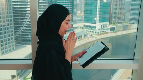 الانجاز وتحقيق الاهداف، ايماءات وجه ويدين تدل على النجاح والفوز، تصوير مقرب من الجانب لامراة عربيه خليجيه اماراتية ترتدي العباءة و الحجاب تقف بجانب النافذة و تمسك بيديها التابلت، تعابير الدهشه و السرور، سيده سعوديه تقوم بادارة مهام العمل اليومية، استخدام الاجهزة التقنية الحديثة في العمل