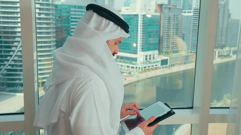النقر على شاشة الجهاز اللوحي، النظر الى شيء ما ، ادارة مهام العمل اليومية، استخدام الاجهزة التقنية الحديثة في العمل، تصوير من الجانب لرجل عربي خليجي اماراتي يرتدي الكندورة و الغترة يقف بجانب النافذة يحمل بيده جهاز التابلت
