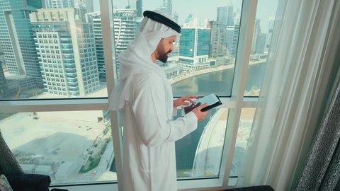 النظر الى شيء ما ، ادارة مهام العمل اليومية، استخدام الاجهزة التقنية الحديثة في العمل، تصوير من الجانب لرجل عربي خليجي اماراتي يرتدي الكندورة و الغترة يقف بجانب النافذة يحمل بيده جهاز التابلت