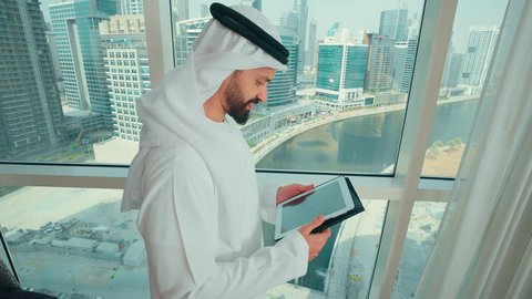 استخدام الاجهزة التقنية الحديثة في العمل، الانجاز وتحقيق الاهداف ، ادارة مهام العمل اليومية، تصوير من الجانب لرجل عربي خليجي اماراتي مبتسم يرتدي الكندورة و الغترة يقف بجانب النافذة يحمل بيده جهاز التابلت،  ايماءات وجه ويدين تدل على النجاح والفوز