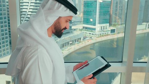 Achievement and goal attainment, managing daily work tasks, using modern technological devices at work, a side shot of a smiling Emirati Gulf Arab man wearing a kandura and ghutrah standing by the window holding a tablet in his hand, facial and hand gestures indicating success and victory.