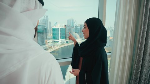 Exchanging chats and conversations at home, the concept of intellectual bonding and understanding between spouses, an Emirati Gulf Arab man wearing a kandura and ghutrah talking to his wife, an Emirati woman in a hijab wearing an abaya pointing to something, gestures of happiness and joy.