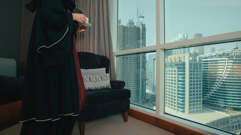 Spending a pleasant and quiet time, vitality and energy when drinking caffeine, a smiling Emirati Gulf Arab woman wearing a black abaya sitting on a chair by the window enjoying hot beverages, a Saudi woman holding a coffee cup in her hands.