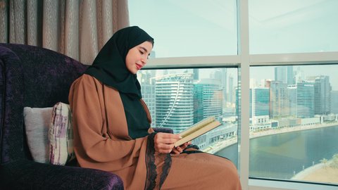 قضاء وقت الفراغ بالتعلم والثقافة، ثقافة التعلم الذاتي، الاستمتاع بقراءة الكتب والقصص والروايات، سيده سعودية تقوم بممارسة هواية القراءة والمطالعة، امراة عربية خليجية اماراتية محجبة ترتدي العباءة تجلس على كرسي مريح تحمل بيدها كتاب