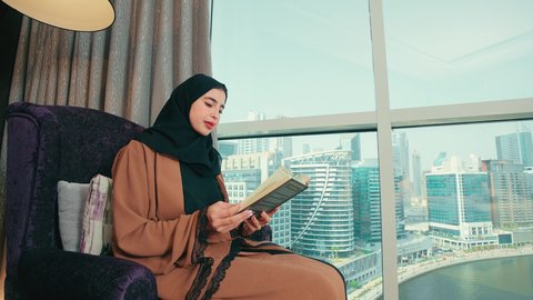 ثقافة التعلم الذاتي، الاستمتاع بقراءة الكتب والقصص والروايات، سيده سعودية تقوم بممارسة هواية القراءة والمطالعة، قضاء وقت الفراغ بالتعلم والثقافة، امراة عربية خليجية اماراتية محجبة ترتدي العباءة تجلس على كرسي مريح تحمل بيدها كتاب