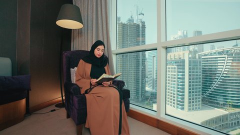 الاستمتاع بقراءة الكتب والقصص والروايات, ثقافة التعلم الذاتي،سيده سعودية تقوم بممارسة هواية القراءة والمطالعة، قضاء وقت الفراغ بالتعلم والثقافة، امراة عربية خليجية اماراتية محجبة ترتدي العباءة تجلس على كرسي مريح تحمل بيدها كتاب