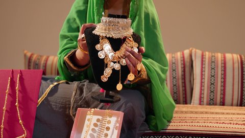 امراة عربيه خليجيه اماراتية ترتدي العباءة المطرزة تحمل بيديها عقد من الذهب لتقديمه للعروس، ذهب محلي اماراتي وطني، العادات والتقاليد الإماراتية في مراسم الزواج، تقديم الهدايا في المناسبات السعيدة