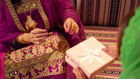 Gift-giving during holidays and happy occasions, Emirati traditions in weddings, a close-up shot of an Emirati Gulf Arab bride wearing a colorful abaya holding a gold necklace in her hands, a Gulf woman presenting a gift box to the bride.