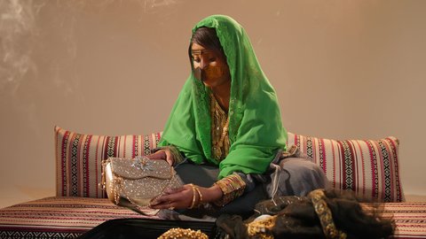 العطاء والاحتفال بالمناسبات السعيدة، الاستعداد لتقديم الهدايا للعروس، مراسم الزواج في المجتمع الإماراتي، امراة عربية خليجية إماراتية ترتدي عباءة مطرزة  تجلس على الاريكة تحمل حقيبة من الذهب