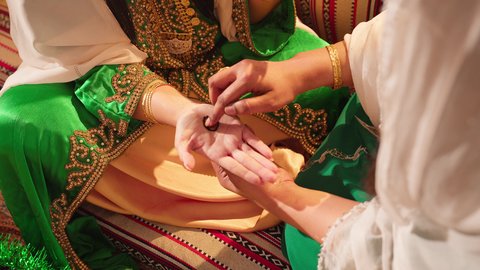 زخارف وفنون يدويه من مواد طبيعيه، عادات وتقاليد في المناسبات والأفراح السعيدة، صورة مقربة ليد إمرأة عربية خليجية إماراتية ترتدي الزي التراثي و تقوم برسم و نقش الحنة على يد شابة، مهن تقليدية شعبية نسائية، التجمل والاهتمام بالمظهر الخارجي على الطريقة الشعبية