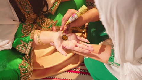 عادات وتقاليد في المناسبات والأفراح السعيدة، صورة مقربة ليد إمرأة عربية خليجية إماراتية ترتدي الزي التراثي و تقوم برسم و نقش الحنة على يد فتاة، مهن تقليدية شعبية نسائية، زخارف وفنون يدويه من مواد طبيعيه، التجمل والاهتمام بالمظهر الخارجي على الطريقة الشعبية