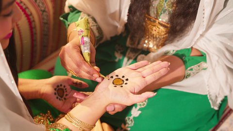 Traditional women's popular fashion, arts, designs, and handmade decorations from natural materials, beautification and decoration, and attention to external appearance, customs and traditions in happy occasions and celebrations, an Emirati Gulf Arab woman is drawing and engraving on the hand of a young Saudi woman using red henna.