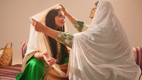 مناسبات وأفراح واحتفالات سعيدة، الاهتمام بالأناقة والمظهر الخارجي، مفهوم التزين والاطلالة المتألقة، سيدة عربية خليجية اماراتية ترتدي الزي التقليدي والبرقع تقوم بوضع الطرحة للعروسة، إيماءات السعادة والفرح ، الاحتفال بليلة العمر