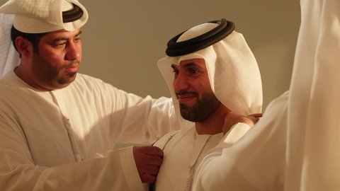 Offering congratulations and assisting in wearing the Emirati bisht, young men celebrating their friend at his wedding, the Al Ayala performance group, a performing art that combines dance, celebrations at Emirati weddings, folk theatrical performances, folk dances at weddings and events, close-up photography to revive wonderful atmospheres for an Emirati Gulf Arab groom.