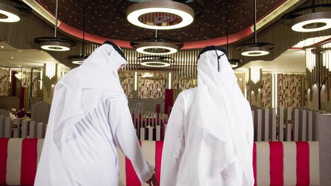 التقدم الى الامام، ايماءات تدل على الحركة والمشي, قضاء وقت ممتع في الخارج, الترفيه والتسلية في العطلة, تصوير من الخلف لرجلان عربيان خليجيان اماراتيان يرتديان الكندورة و الغتره يسيران داخل المطعم, الاستمتاع بتناول وجبات لذيذة