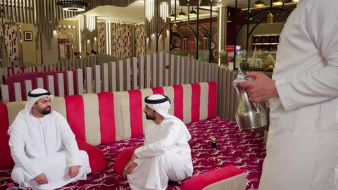 قضاء وقت ممتع في الخارج, رجل عربي خليجي اماراتي يرتدي الكندورة والغترة البيضاء يجلس مع صديقه في مطعم فاخر, الاستمتاع بالمشروبات الساخنة، الترفيه والتسلية في العطلة, تصوير مقرب من الجانب لنادل يقوم  بصب المشروب للزبائن, ايماءات السعادة والسرور