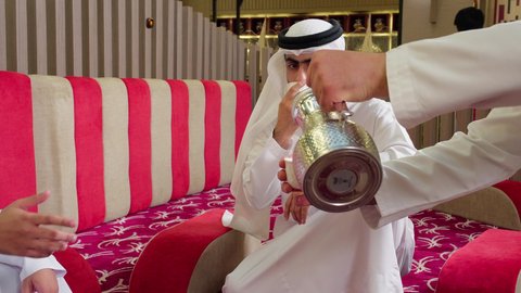 تصوير مقرب لنادل يقوم  بصب المشروب للزبائن, الترفيه والتسلية في العطلة, ايماءات السعادة والسرور، مشاركة المشروبات الساخنة والقهوة، قضاء وقت ممتع في الخارج، ،رجل عربي خليجي اماراتي يرتدي الكندورة والغترة البيضاء يجلس مع صديقه في مطعم فاخر