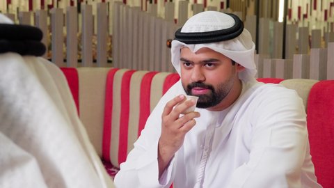 قضاء وقت ممتع في الخارج,  مشاركة المشروبات الساخنة والقهوة, ايماءات السعادة والسرور، تبادل الاحاديث بين الأصدقاء،تصوير مقرب لرجل عربي خليجي اماراتي يرتدي الكندورة والغترة البيضاء يجلس مع صديقه في مطعم فاخر