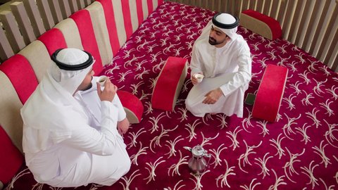 ايماءات السعادة والسرور، مشاركة المشروبات الساخنة والقهوة، قضاء وقت ممتع في الخارج، تبادل الاحاديث بين الأصدقاء،رجل عربي خليجي اماراتي يرتدي الكندورة والغترة البيضاء يجلس مع صديقه  في مطعم فاخر