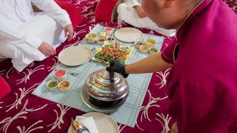 اجتماع الاصدقاء على مائدة مليئة بالاكلات الشعبيه التقليديه الاماراتيه, تصوير مقرب لخادمه تقوم بوضع طبق شعبي على سفرة الطعام
