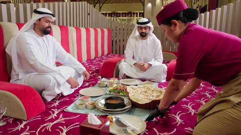 نادلة ترتدي الزي الرسمي تقوم بخدمة الزبائن وتقديم الطلبات لهم , مطاعم و كافيهات في الإمارات ، تناول الغداء في المطعم, تصوير مقرب لرجلان عربيان خليجيان اماراتيان يرتديان الكندورة و الغتره يجلسان على المائدة يقومان بتناول الطعام ، قضاء الأوقات الممتعة في الخارج