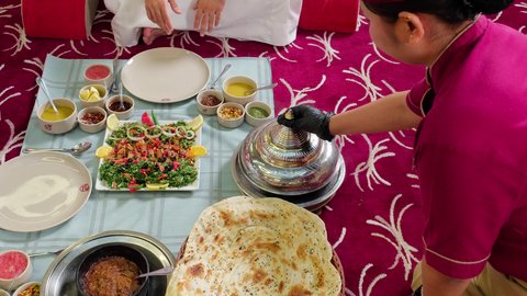Cafes and restaurants in the UAE, unique and special seating, close-up shot of a waitress serving food, two Arab Gulf Emirati friends wearing kandura and ghutrah sitting in a restaurant, enjoying food and having a good time outdoors with friends.