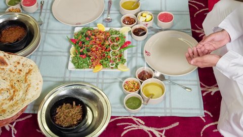 Lunch at the restaurant, gestures of happiness and joy, restaurants and cafes in the UAE, a close-up shot of the hand of an Emirati Arab man in a kandura and ghutrah sitting at the table making a prayer before eating, spending enjoyable times outdoors, a table filled with a variety of delicious Eastern dishes.