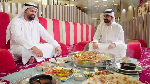 ايماءات السعادة و السرور, مطاعم و كافيهات في الإمارات ، تناول الغداء في المطعم, رجلان عربيان خليجيان اماراتيان يرتديان الكندورة و الغتره يجلسان على المائدة يقومان بتناول الطعام ، قضاء الأوقات الممتعة في الخارج، سفرة مليئة باصناف من المأكولات الشرقية الشهية