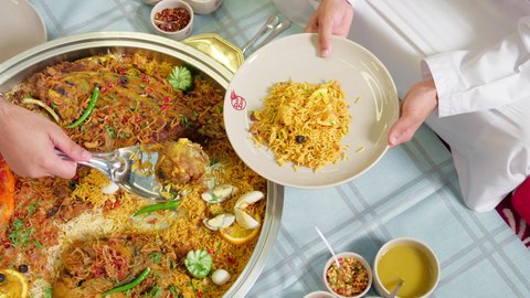 سكب الطعام في الصحون و توزيعه , قضاء أوقات ممتعة, طبق أرز شعبي مع اللحم المبهر مجهز في المطبخ الاماراتي , وصفات الأكلات الشعبية, تصوير مقرب  لرجل عربي خليجي اماراتي يرتدي الكندورة يجلس على سفرة الطعام  يقوم بتحضير طبق الطعام