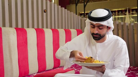 Recipes for traditional dishes, restaurants and kitchens of the United Arab Emirates, an Arab Gulf Emirati man wearing a kandura and ghutrah sitting at the dining table in the restaurant having lunch.