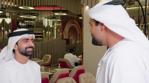Gestures of welcome and peace, a session of youth entertainment and fun, friendship and acquaintance, and building relationships. Two young Arab Gulf Emiratis wearing kandura and ghutrah are sitting at a table in an upscale restaurant, enjoying a good time outdoors.