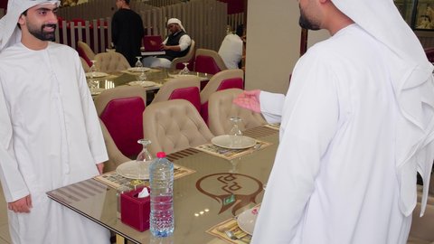جلسة تسلية وترفيه شبابية، صداقة وتعارف وبناء علاقات، ايماءات الترحيب و السلام، شابان عربيان خليجيان اماراتيان يرتديان الكندورة و الغتره يجلسان في مطعم راقي أمام طاولة ، قضاء وقت ممتع في الخارج
